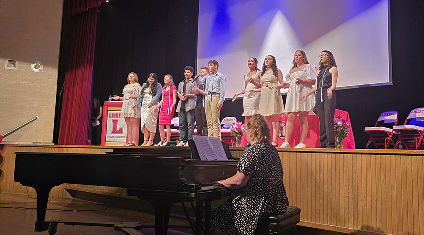Students sing on stage as a woman plays piano in front of the stage