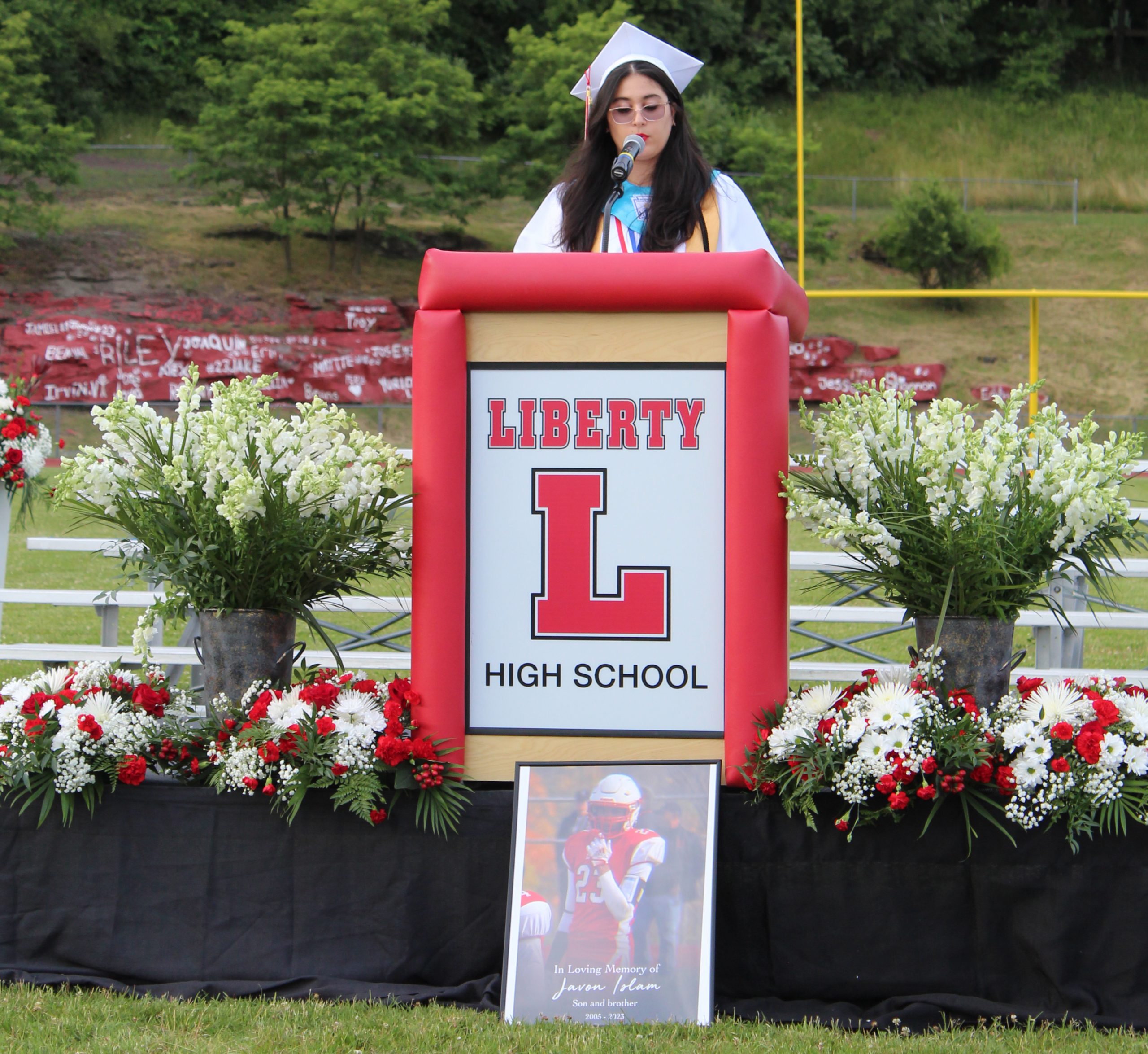 A graduate speaks at a podium with a framed photo of a classmate sitting below the podium.