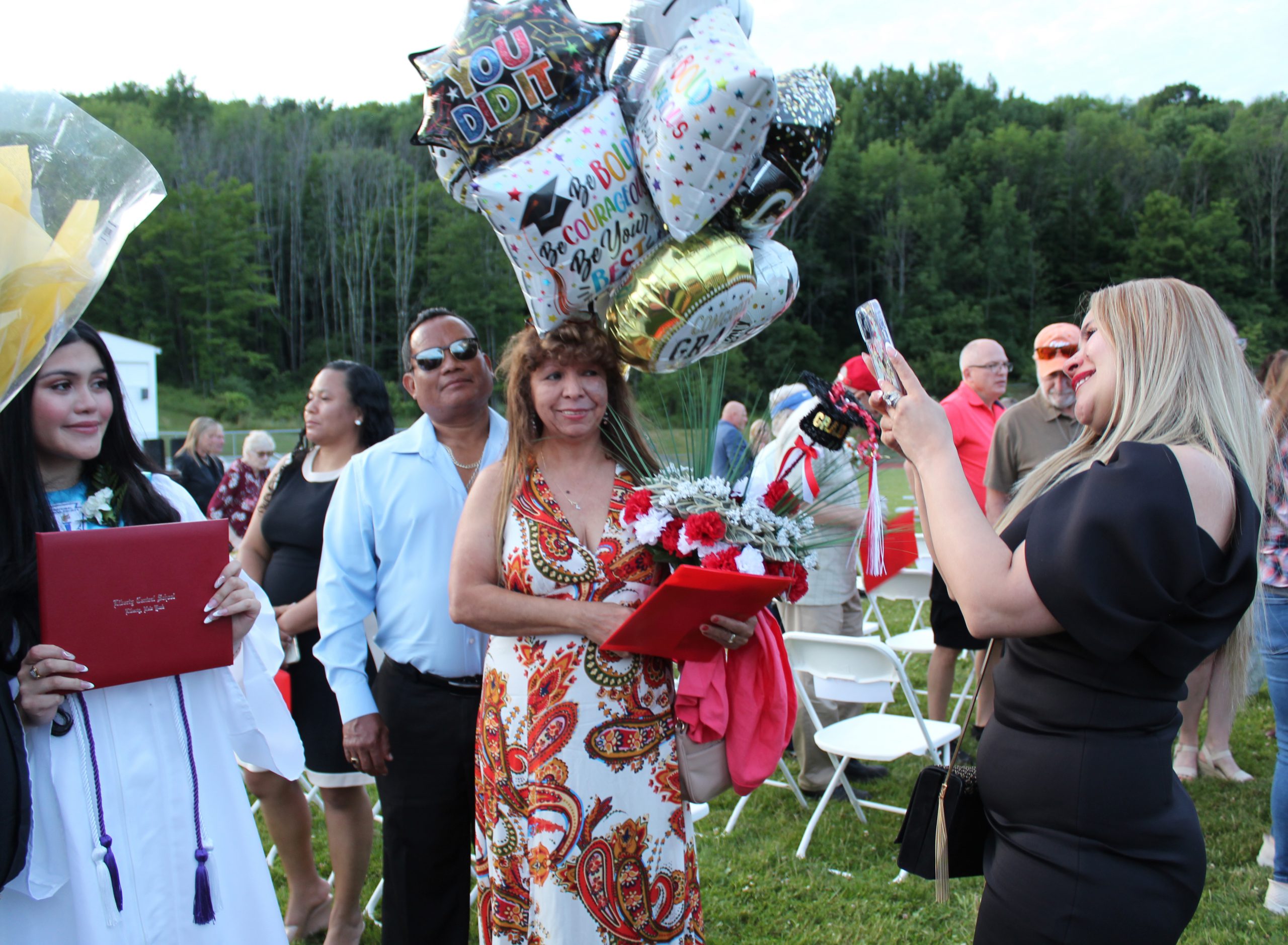 An adult takes a photo of a graduate as another adult holds a bunch of balloons