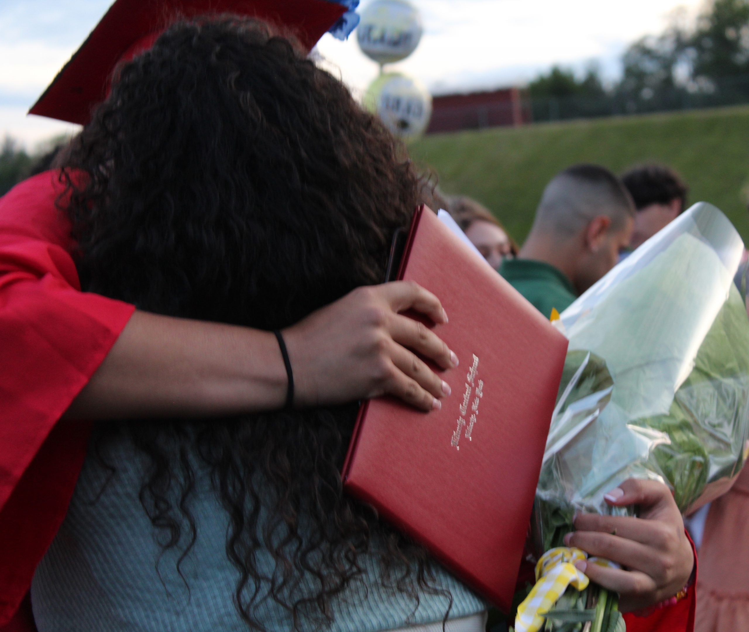 A graduate holding a diploma and flowers hugs an adult