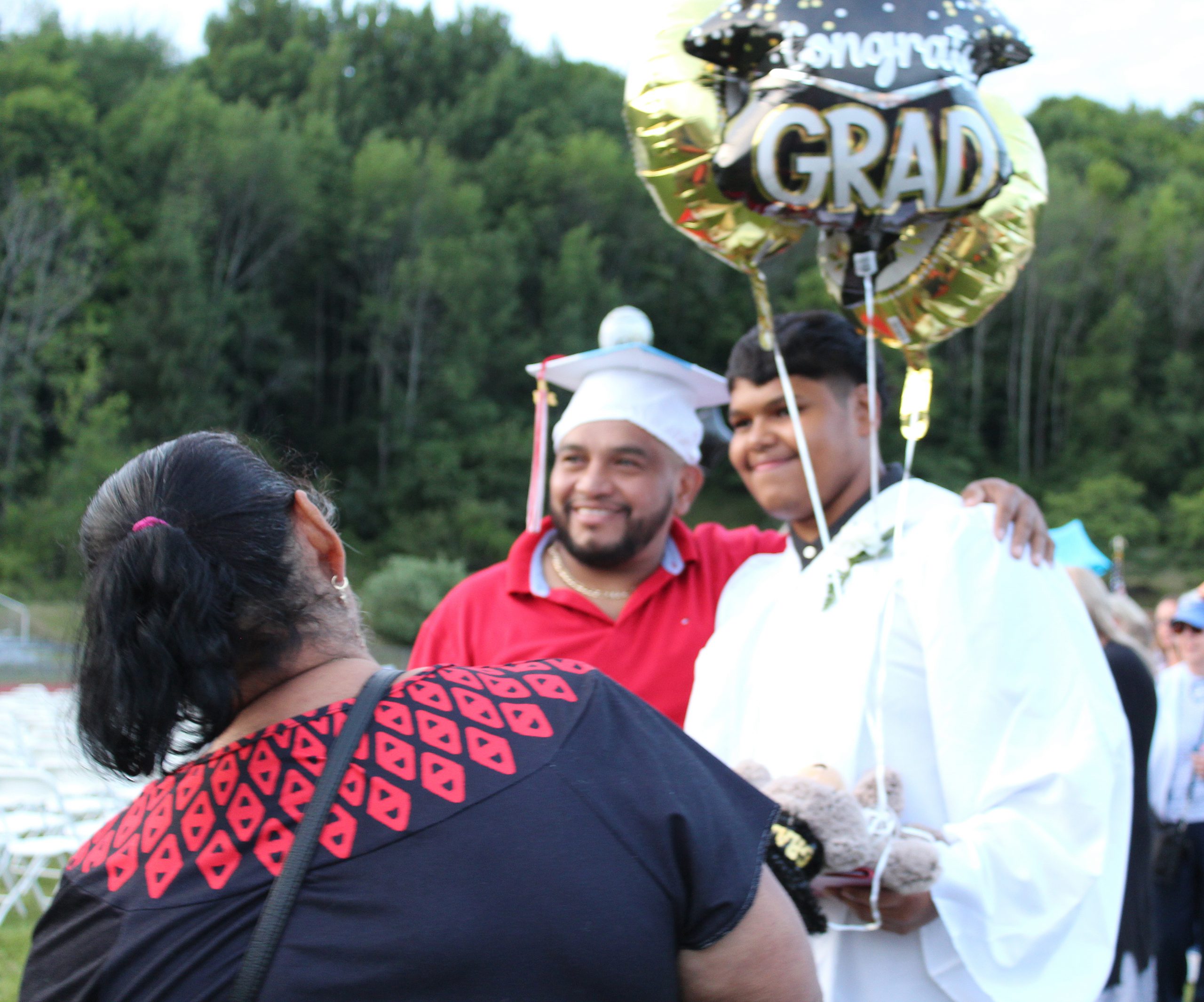 A woman watches as a man poses with a student in a gown holding balloons as the man wears the cap