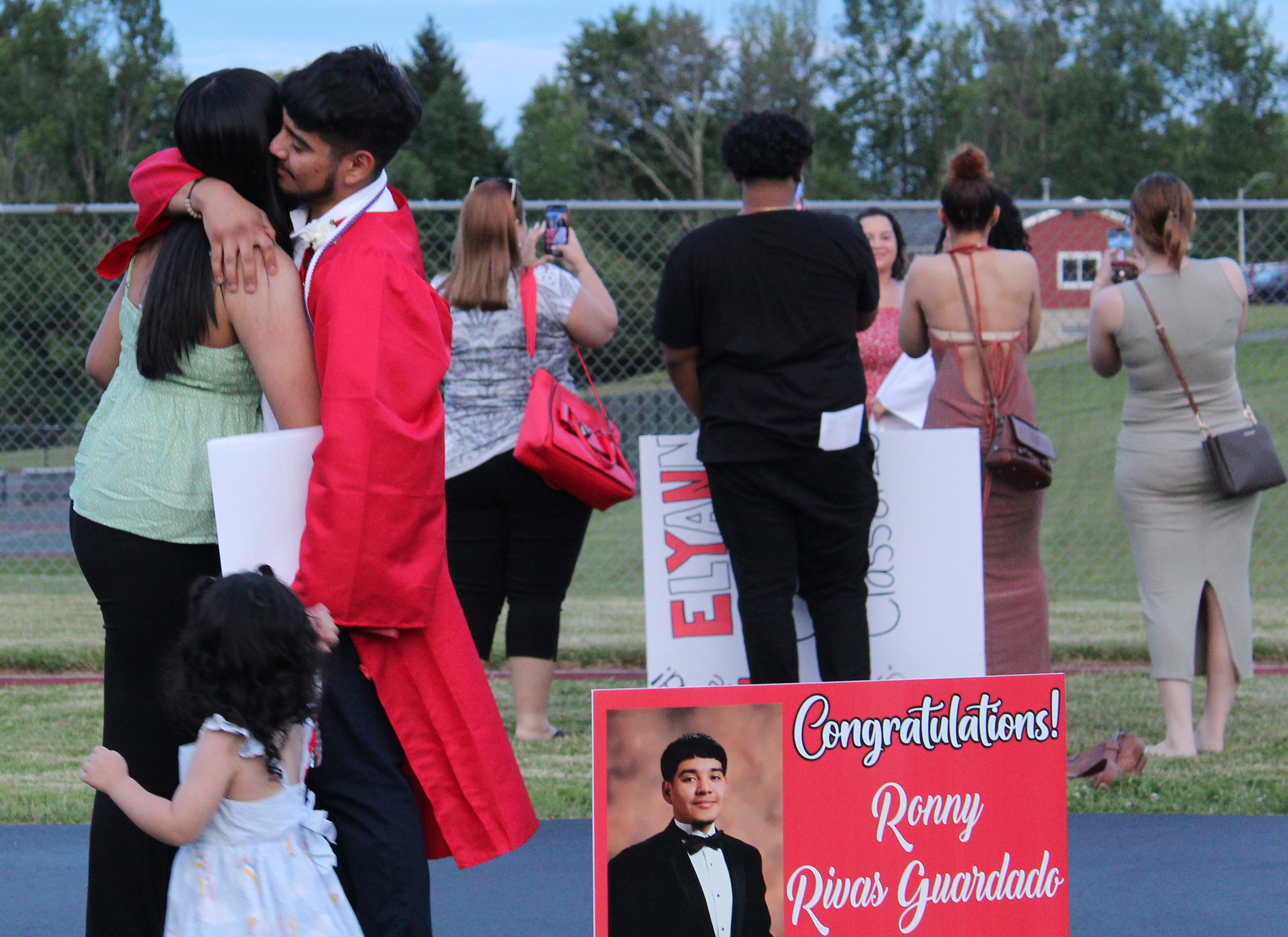 A graduate hugs an adult with a sign graduation sing in front of them as another group prepares to take photos in the back ground