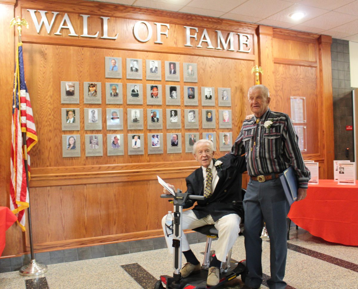 A man stands next to a man seated on a scooter in front of a wall featuring plaques.