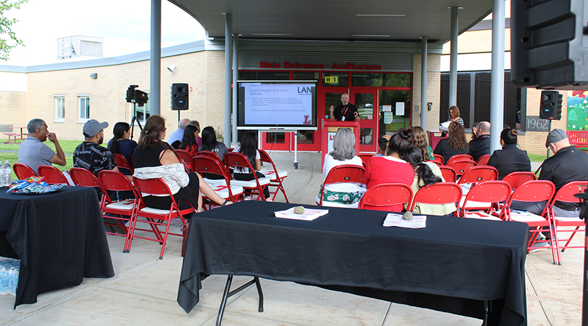 A man speaks at a podium in front of a large screen as members of the audience sit in red folding chairs