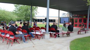 A woman speaks seated at right as others sit near her and next to the audience in red folding chairs as a woman stands at a podium