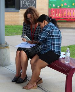 Two woman look at a phone while seated on a bench.