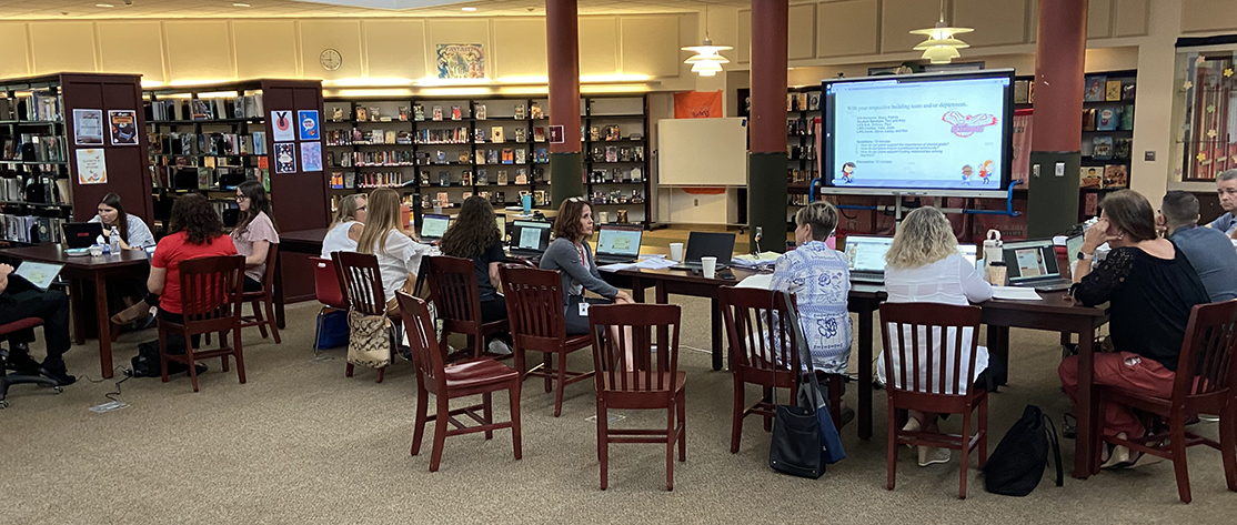 People talk at tables in a library as a large tv screen shows a slide from a presentation.