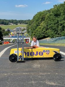 A student poses with her yellow soap box derby car with Mojo written on the side and a trophy standing next to it.