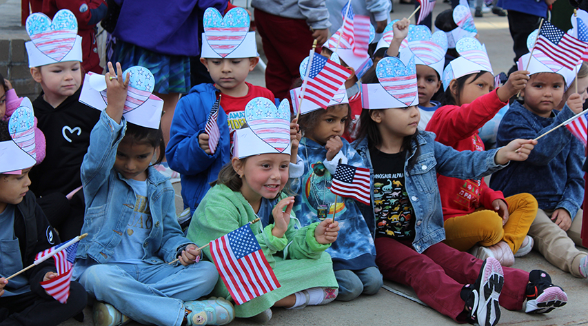 Students wearing paper hats in red white in blue and holding flags sit on the ground as one student in the middle waves.