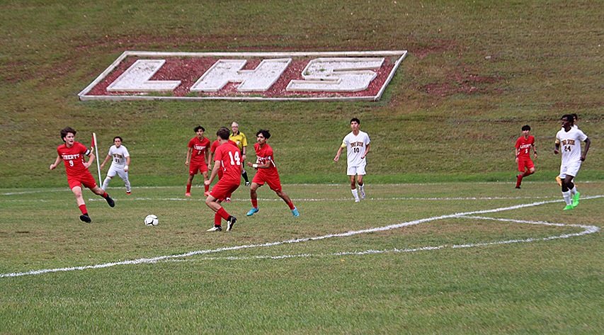 Soccer players in red head toward the ball as players in white lag behind and a ref in yellow watches with the LHS sign in the background