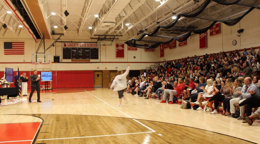 A person waves to the audience a she walks across the gym and three people at left clap