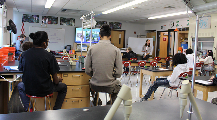 A teacher sits at the front of the class speaking as students sit at tables