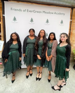 Five students in green dresses and black shoes pose in front of a banner that says Friends of EverGreen Meadow Academy