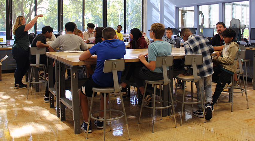 An adult points as students and another adult sit at two high tables