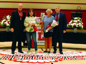 Two man hold flowers flanked by two men behind the Liberty Redhawk logo on the gym floor.