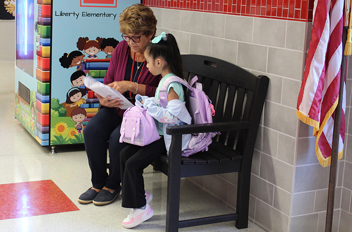 Sitting on a bench, an adult points to a piece of paper sitting next to a young student with a back pack and bag