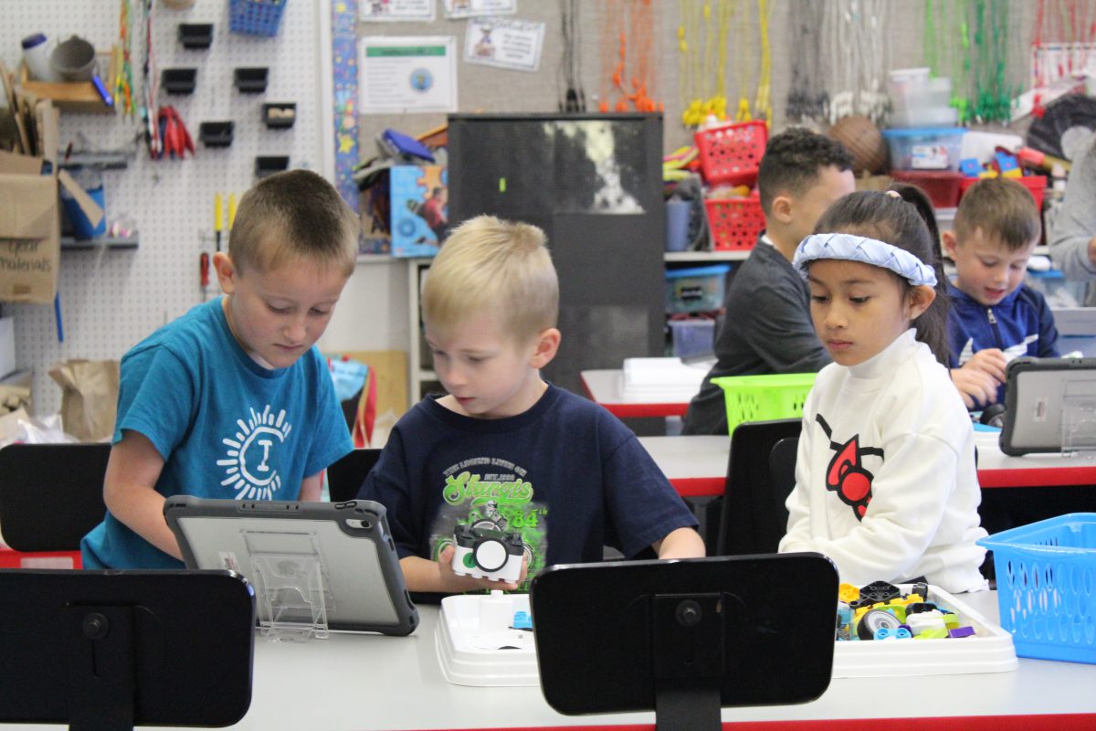 Two students look at the screen of a tablet as another student works on a robot to their left.