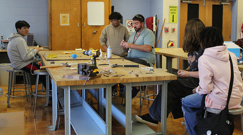 A teacher at a large wooden table demonstrates the project as students watch