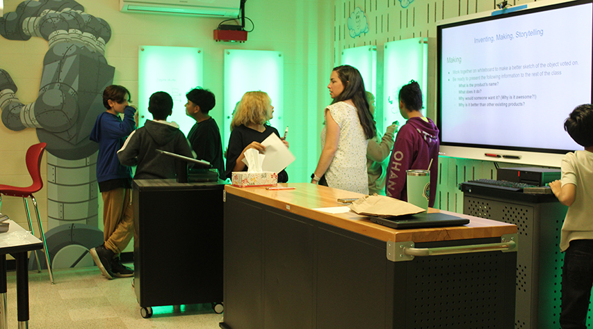 Students work at light up white boards with a screen telling them instructions at right, as an adult talks to a student near a table