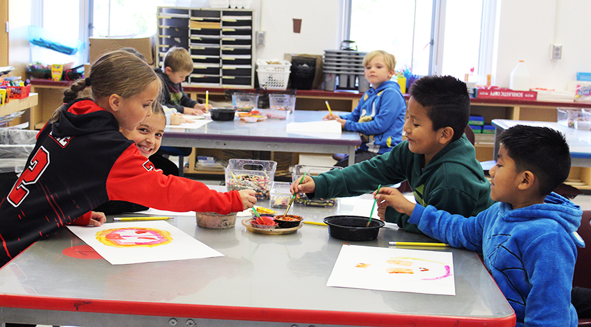 A student smiles behind another student as four students work on paintings at a table and other students work at a desk behind them.