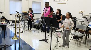 Three students practice drumming on drum pads while standing as an instructor leads them.