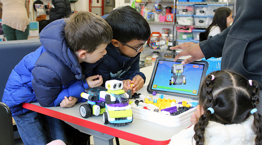 An adult adjusts the screen on a tablet as three students watch as they build a robot.