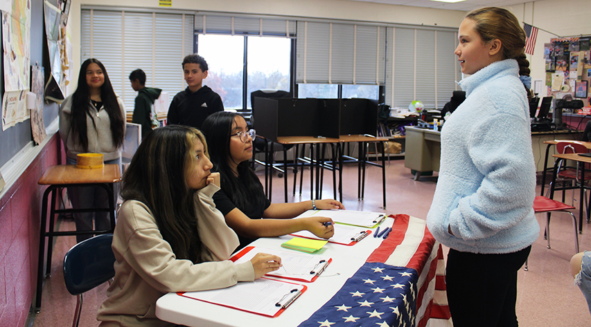 Two students behind a table with an American flag with clipboards ask a student questions as other students stand behind them.