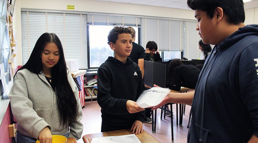 A student standing behind a desk hands a ballot to another student as another student places a card in a bucket.