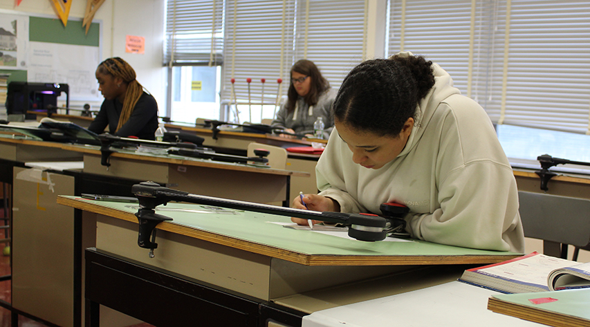 A student works on a drawing at a drafting table as two other students work behind her.