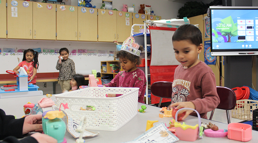 Students play with small toys on a table as students in the background play in a play kitchen.