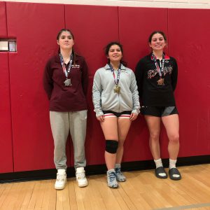 Three girls pose with wearing medals against red padded walls in a gym.