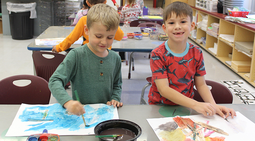 A student looks up to smile from a multi-media artwork as another student paints next to him