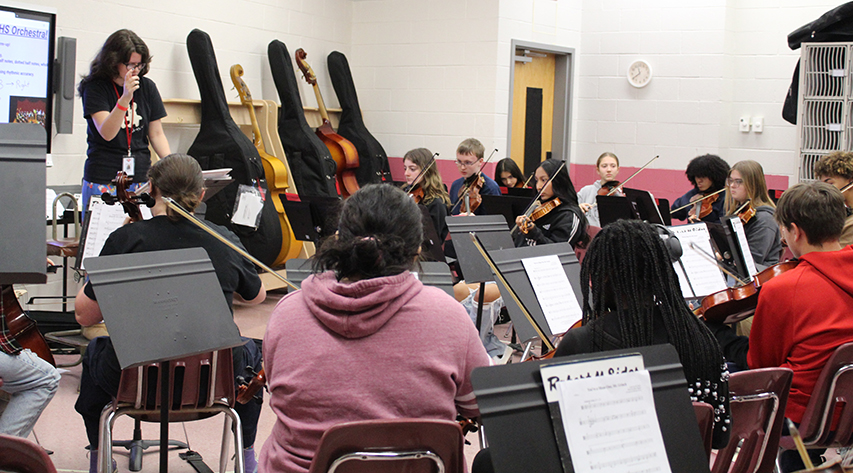 An orchestra teacher conducts students in a band room.