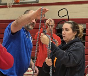 An instructor holds up the end of a rope as a teacher works on tying it properly as another teacher works on a knot in the background.