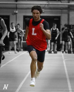 A student in a red shirt with a white L runs down the track