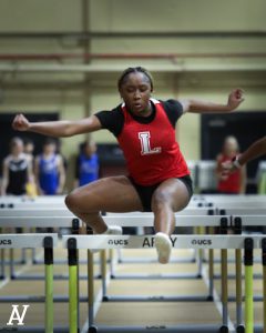 A student in a red shirt with a white L jumps a hurdle