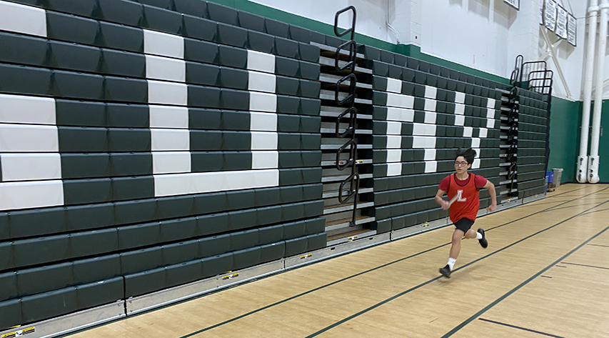 A student in a Liberty uniform runs past green bleachers with SUNY in white letters on them.