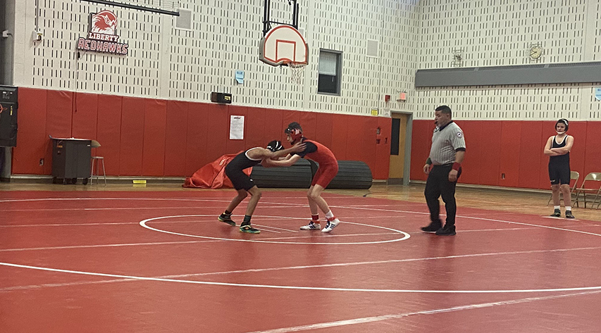 Two wrestlers grapple in the middle of a mat as a referee officiates and another wrestler watches.